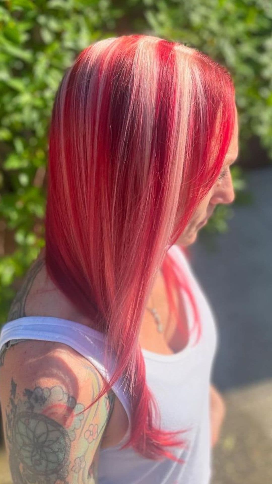 Person with Candy Cane Stripes hairstyle. Shoulder-length hair has red and white stripes. They wear a white tank top and have tattoos on their arm. Background shows greenery.