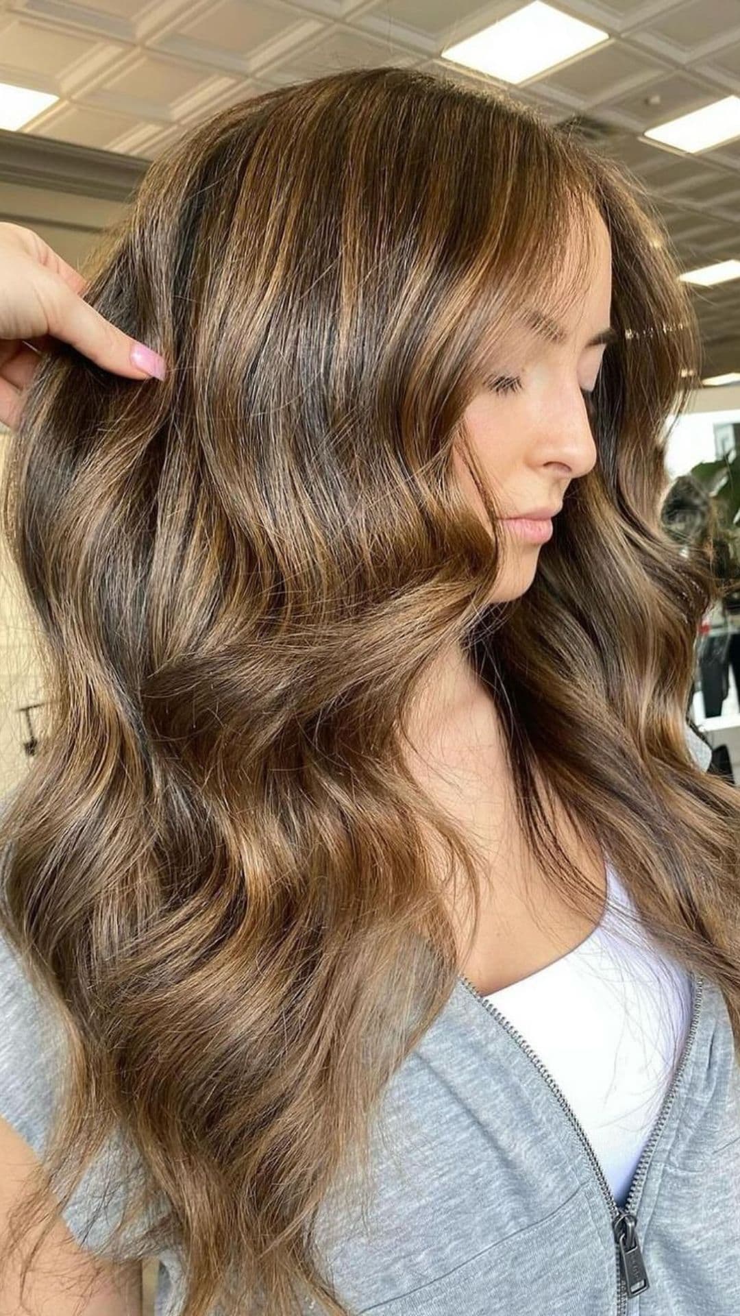 Person showcases long, wavy hair with golden glitter highlights. Casual attire with a gray zip-up hoodie in a well-lit salon.