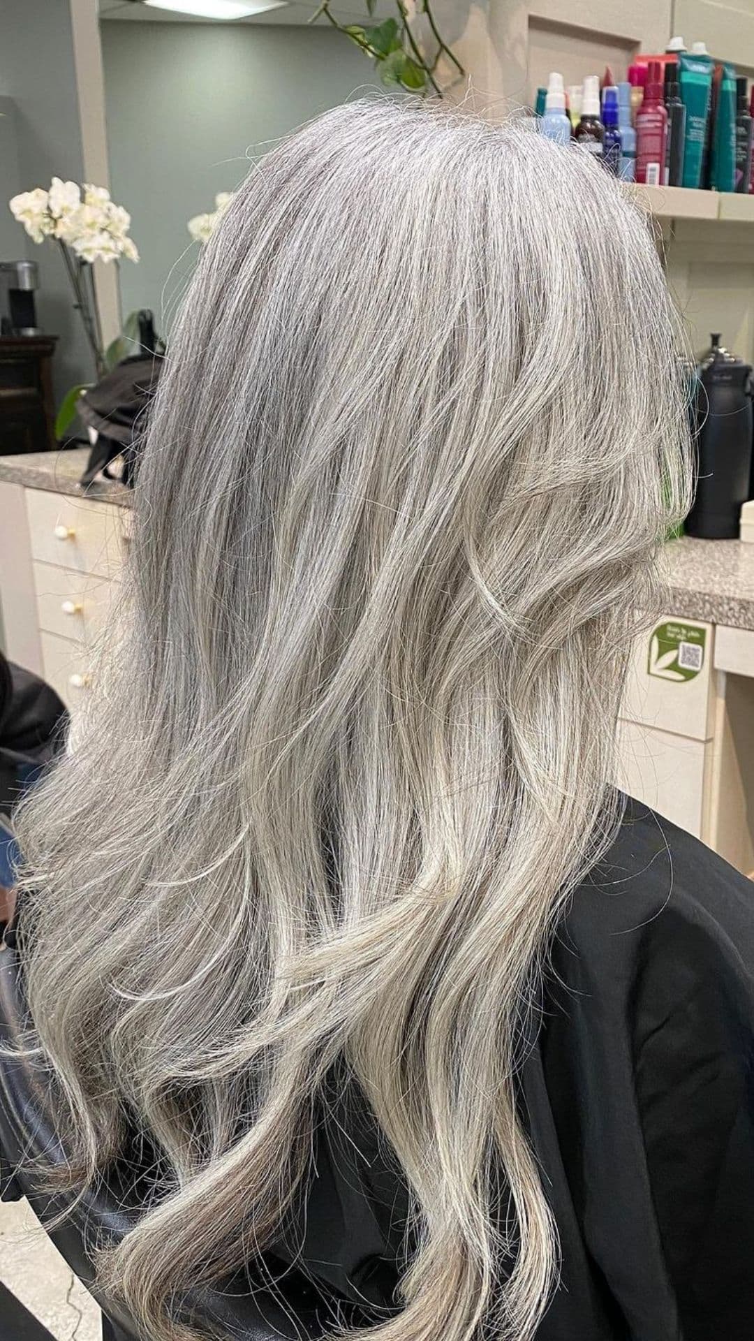 Close-up of a person modeling the Snow Queen Silver hairstyle in a salon setting. The long, wavy silver hair flows smoothly down the back.