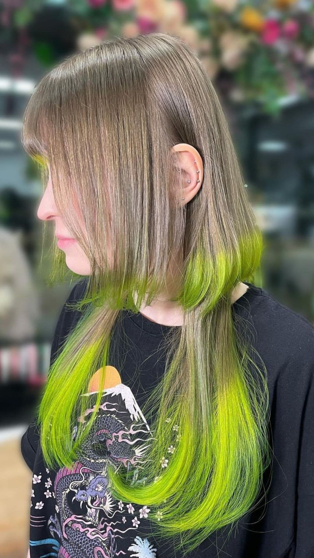 Person with Neon Jellyfish Glow hairstyle, featuring bright green highlights on straight brown hair. Facing left in a casual black shirt with detailed print.