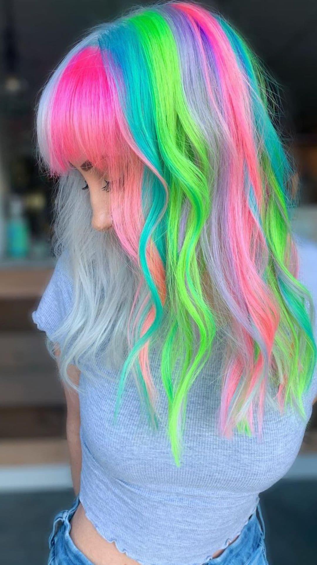 A person with vibrant Pastel Mermaid Hair, featuring pink, blue, green, and purple hues. They're wearing a light gray shirt and looking down. The style showcases a mix of soft colors and waves.