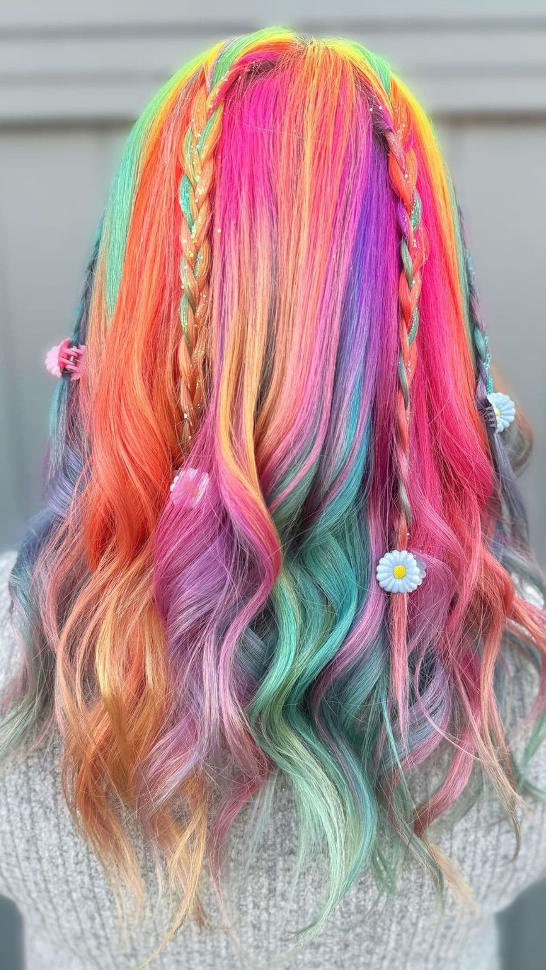 A person models a Rainbow Braids hairstyle. The hair features vibrant colors with two small braids on top. Flowers are woven into the braids for added detail.