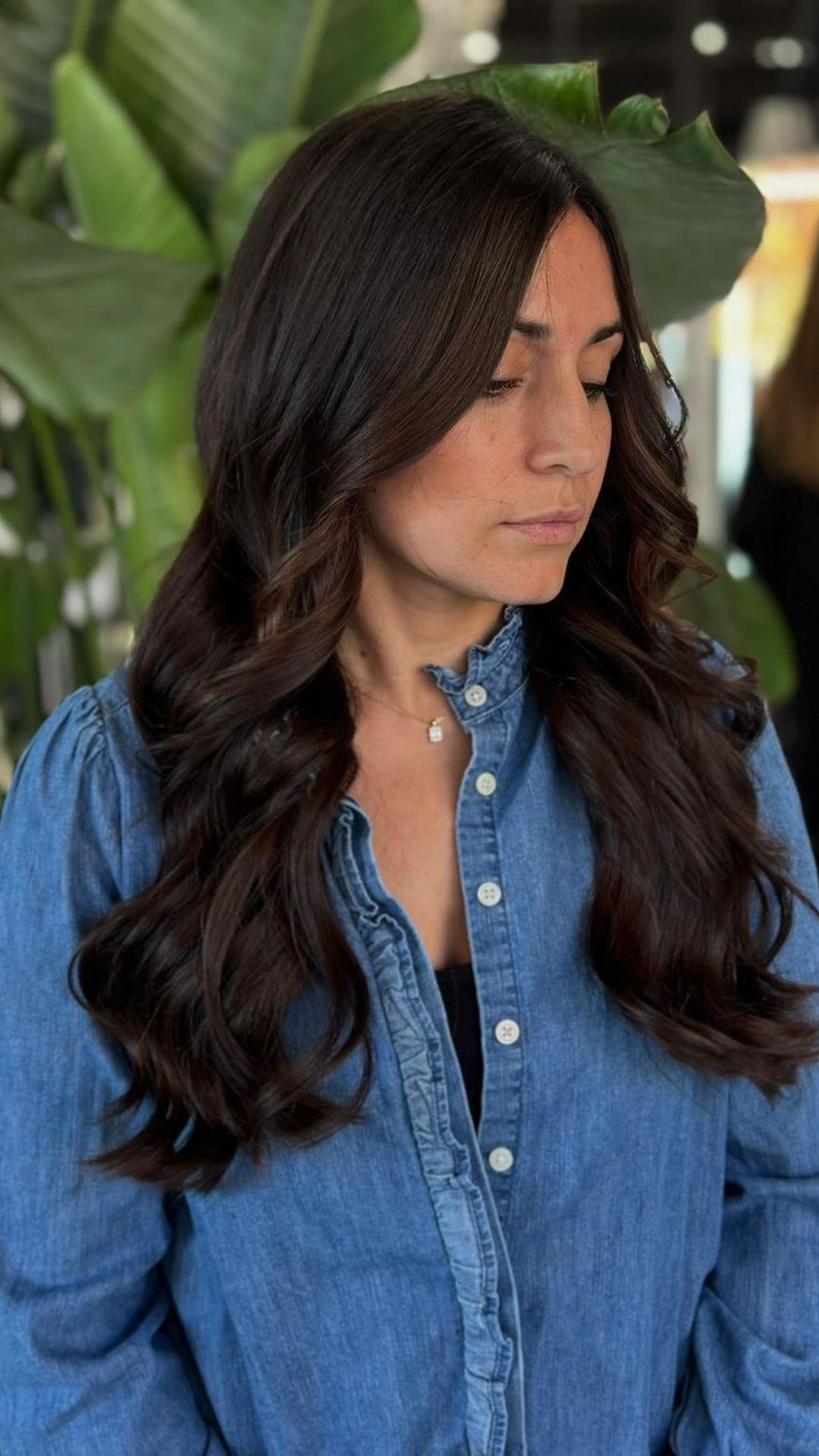 A person with long, wavy chocolate-colored hair wears a denim shirt. They stand in front of green plants, showcasing the hairstyle.