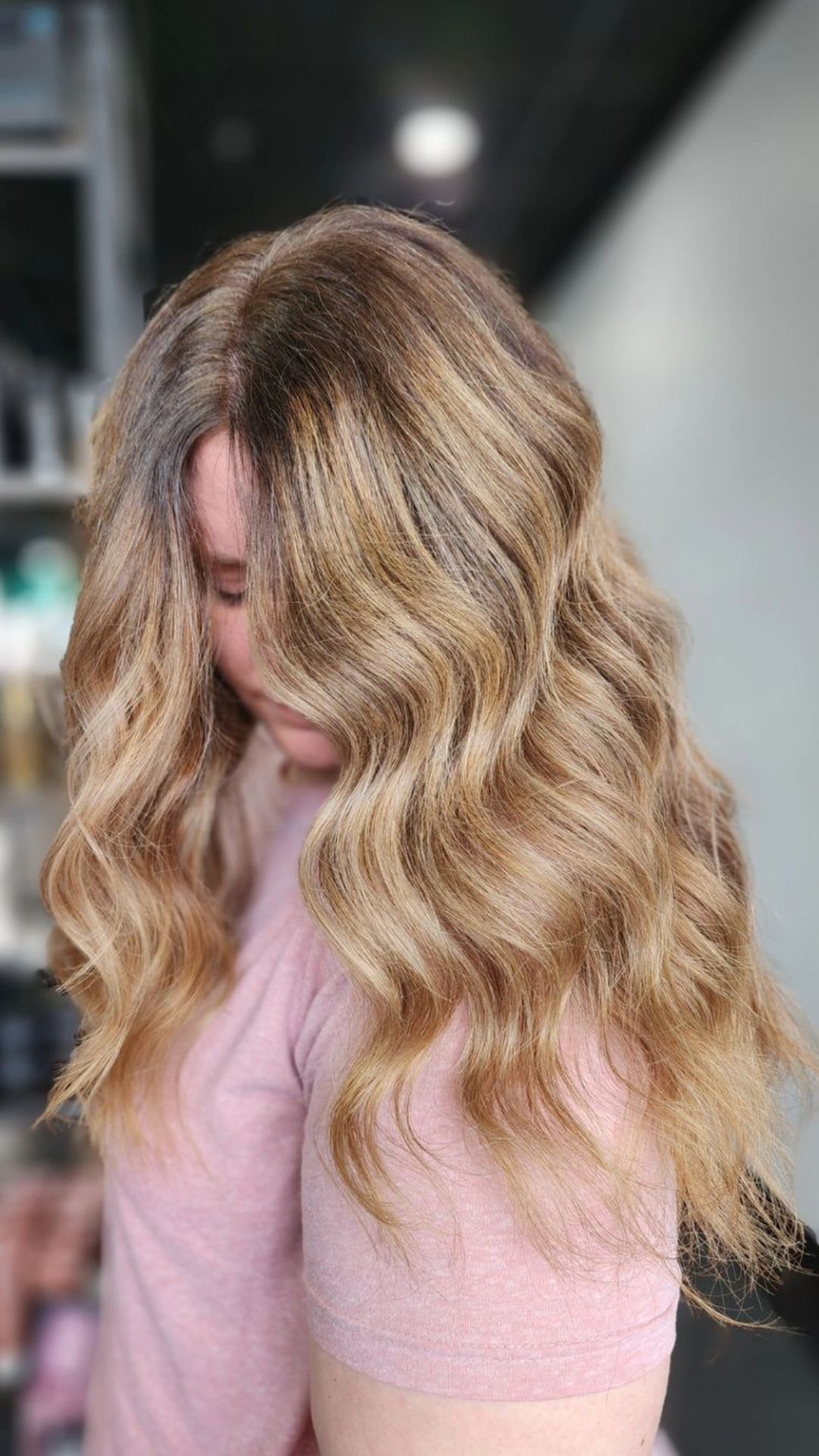 Person with long, wavy golden bronde hair, wearing a pink shirt, tilts head slightly down. The hairstyle features soft, flowing waves.