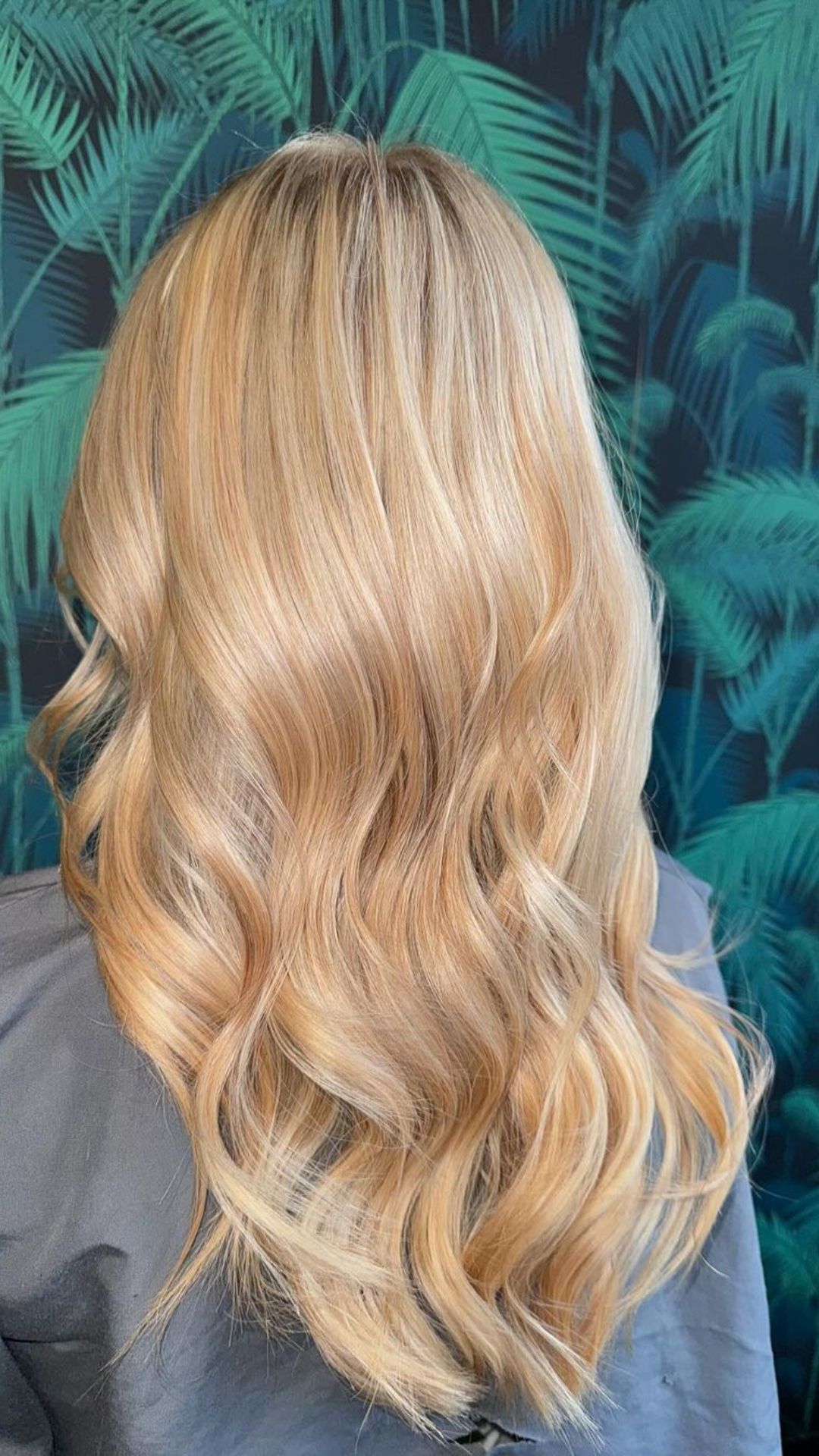 Close-up of a person’s back showcasing a Honey Blonde hairstyle with soft waves against a tropical leaf background.