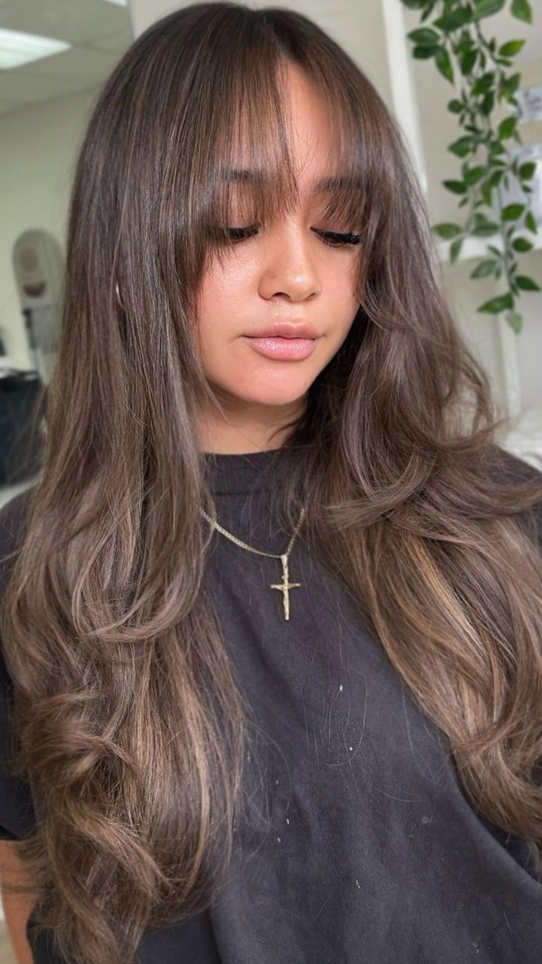 Woman with long, wavy Mushroom Brown hair, wearing a black top and gold necklace.