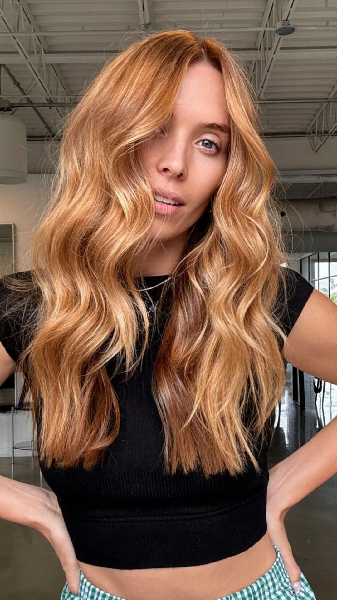 Woman with long, wavy strawberry blonde hair, wearing a black top. She stands in a modern, industrial-style room.