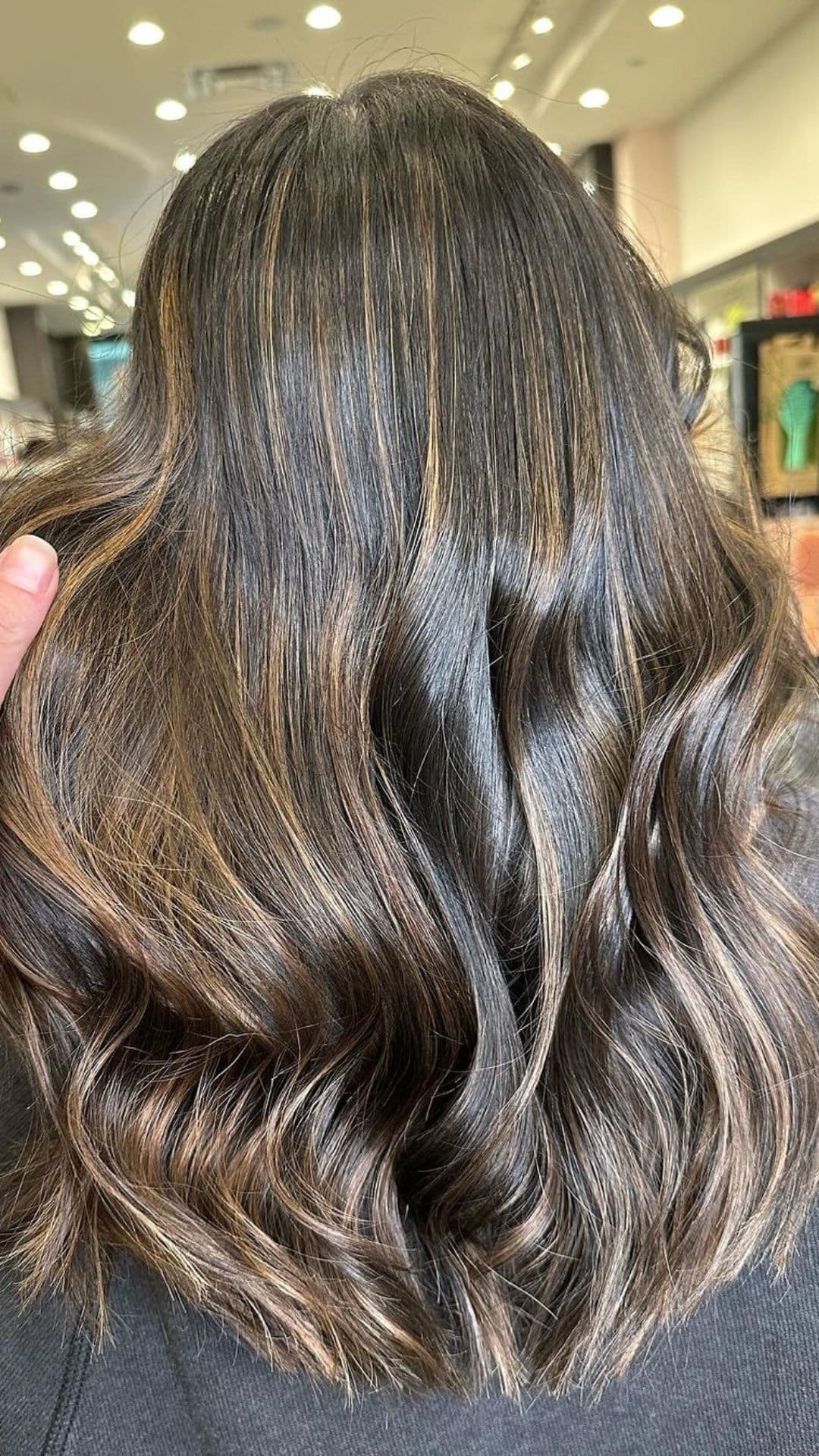 Person with long wavy hair showcasing a Butterscotch Balayage hairstyle. The hair features warm caramel and honey tones, adding dimension and depth. Bright indoor lighting highlights the colors.