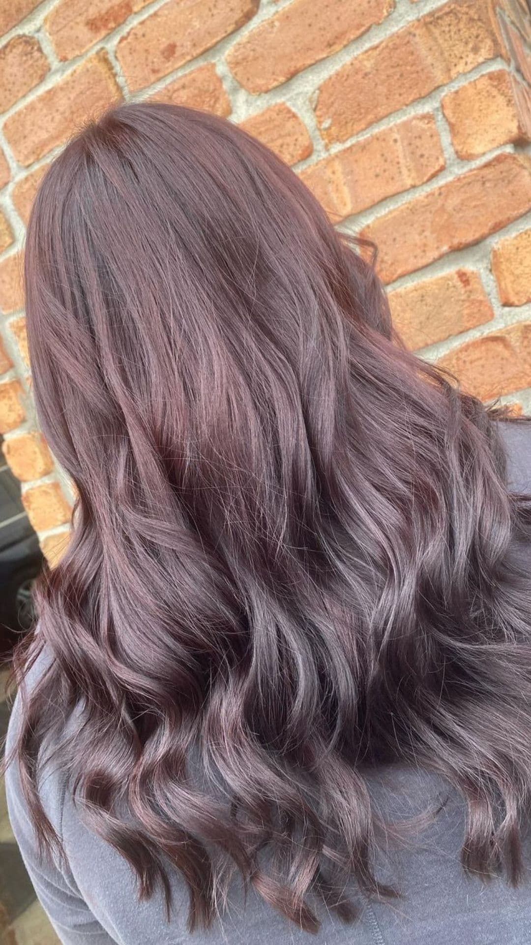 Woman with long, wavy hair featuring a Chocolate Cherry hue, standing in front of a brick wall.