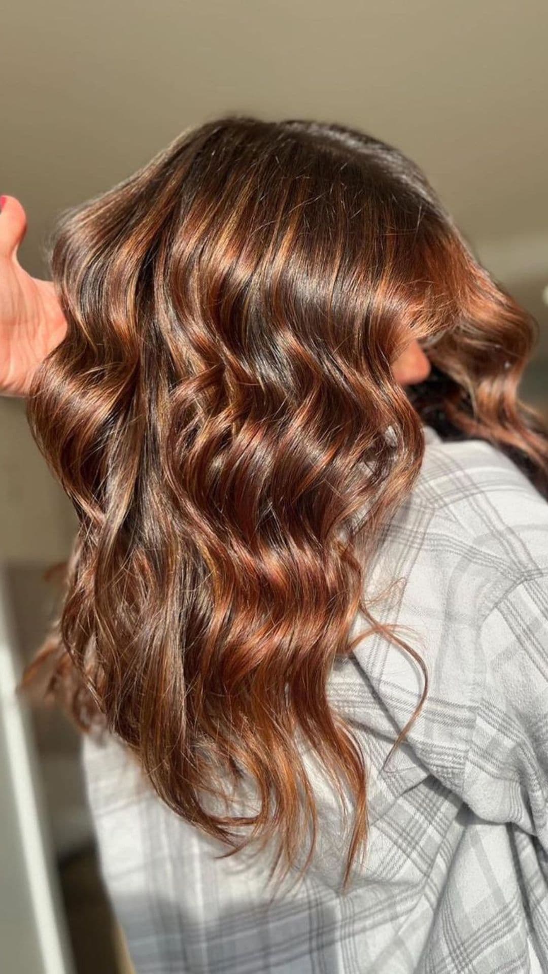 Person wearing a plaid shirt showcasing a Cinnamon Swirl hairstyle. The hair features long, loose waves with warm, blended tones of brown and cinnamon. The style catches the light, highlighting the color variations.