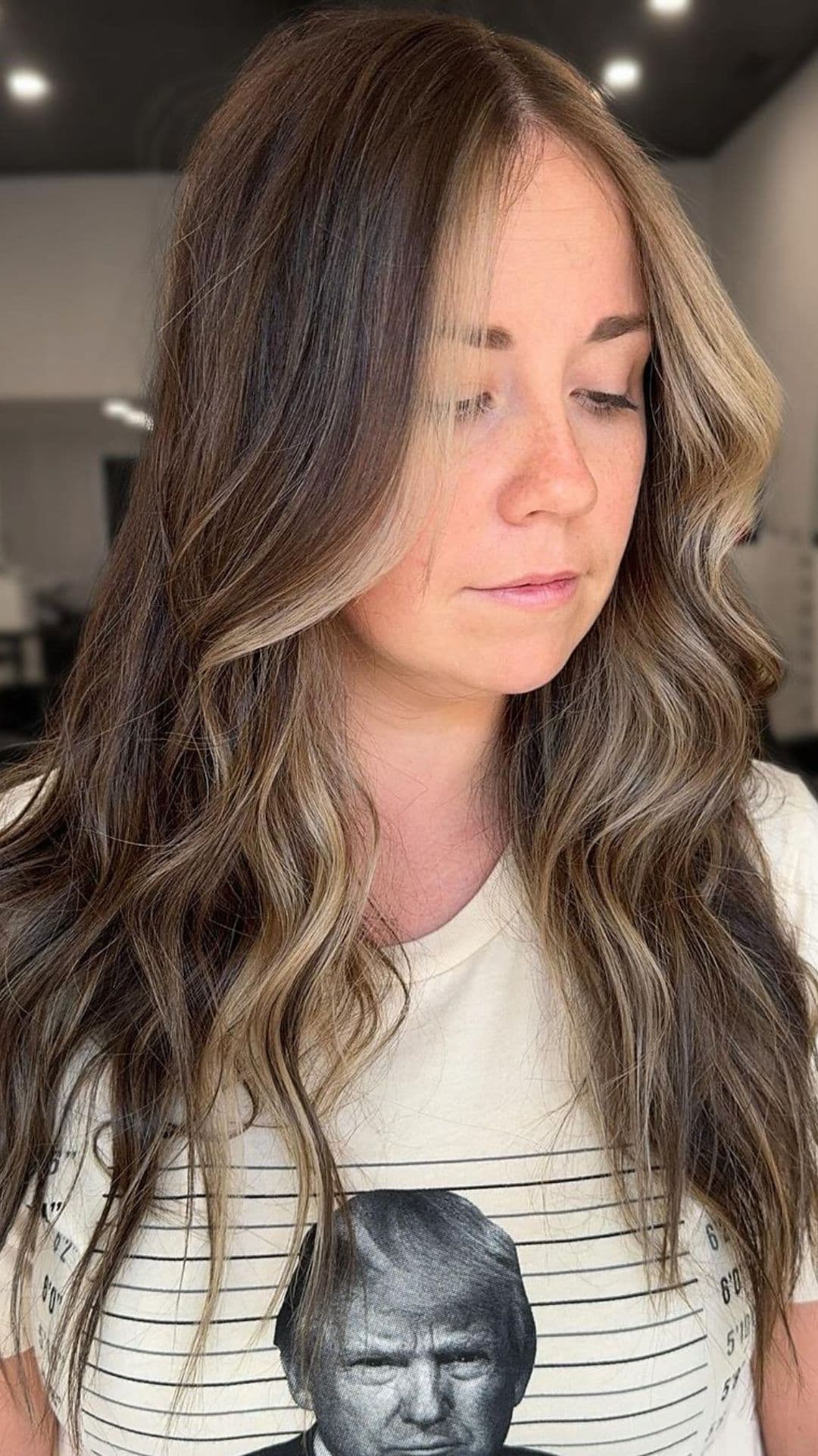 Woman showcasing face-framing blonde highlights hairstyle with wavy brown hair. She’s wearing a graphic t-shirt with a printed photo.