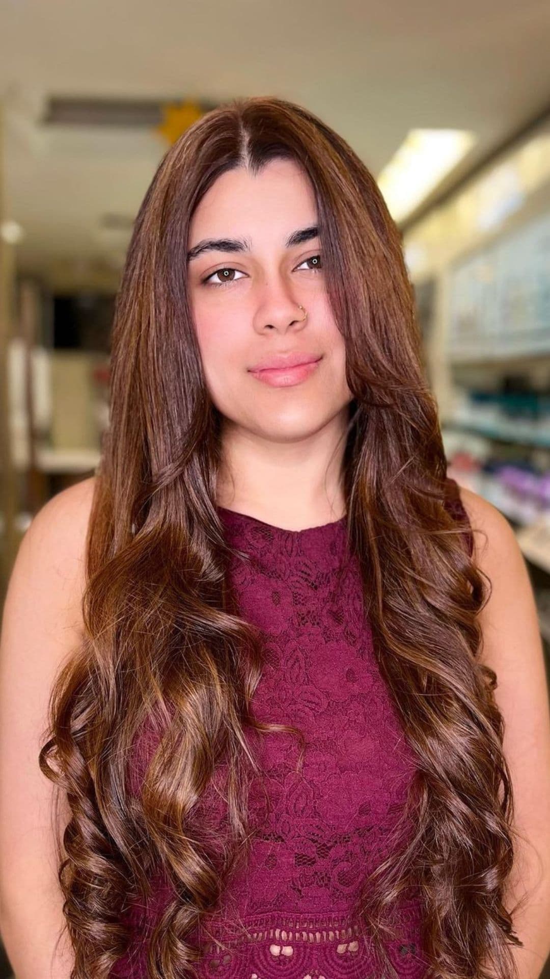 Woman with long, wavy Honey Bronze hair wearing a sleeveless maroon dress in a well-lit room, showcasing a stylish hairstyle.