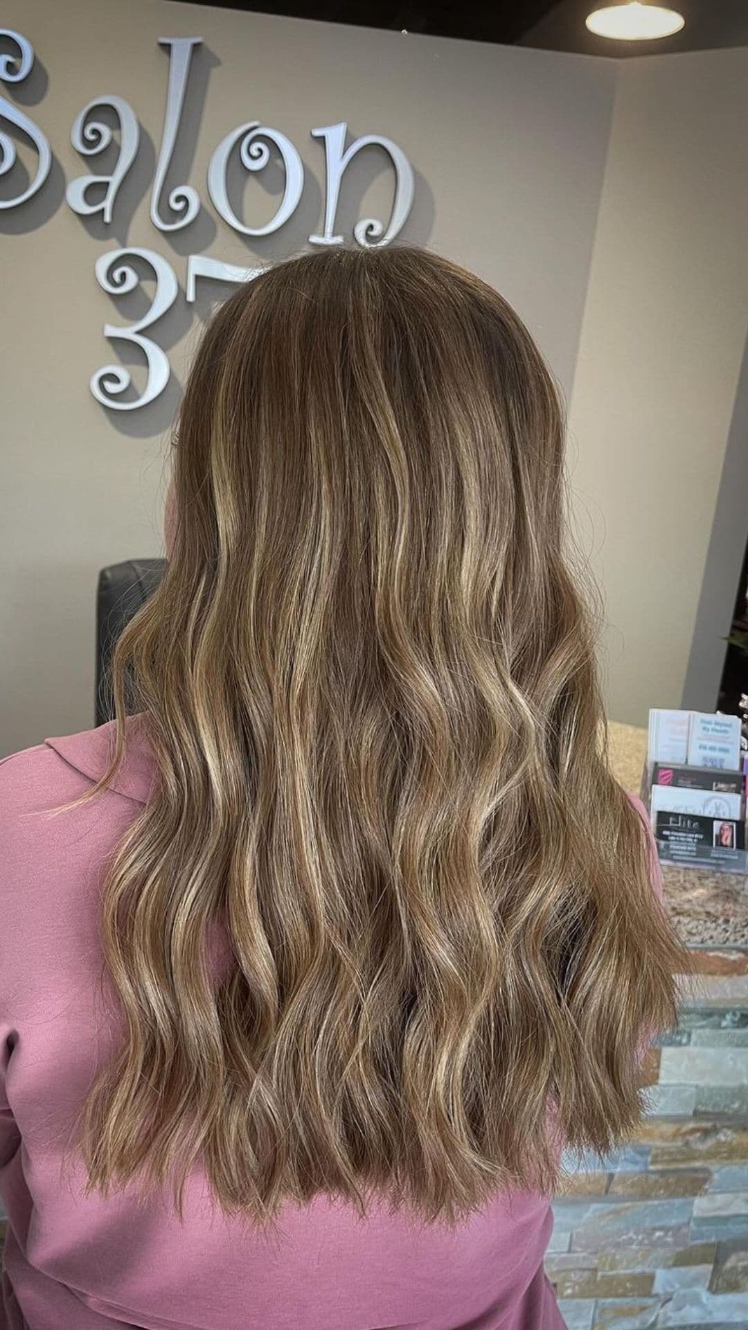 A person with long, wavy hair styled in an Espresso Balayage. They are in Salon 375, wearing a pink top. The background features salon signage and some cards on a counter.