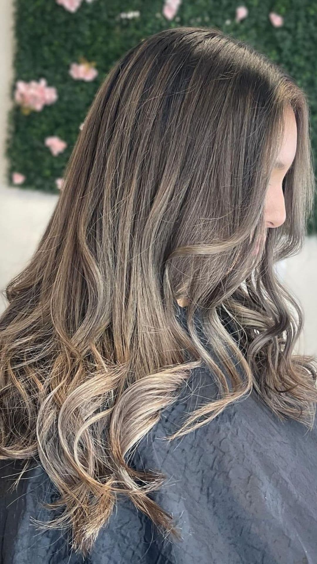 A person with long, wavy hair showcasing an Ashy Brown Balayage hairstyle against a floral background.