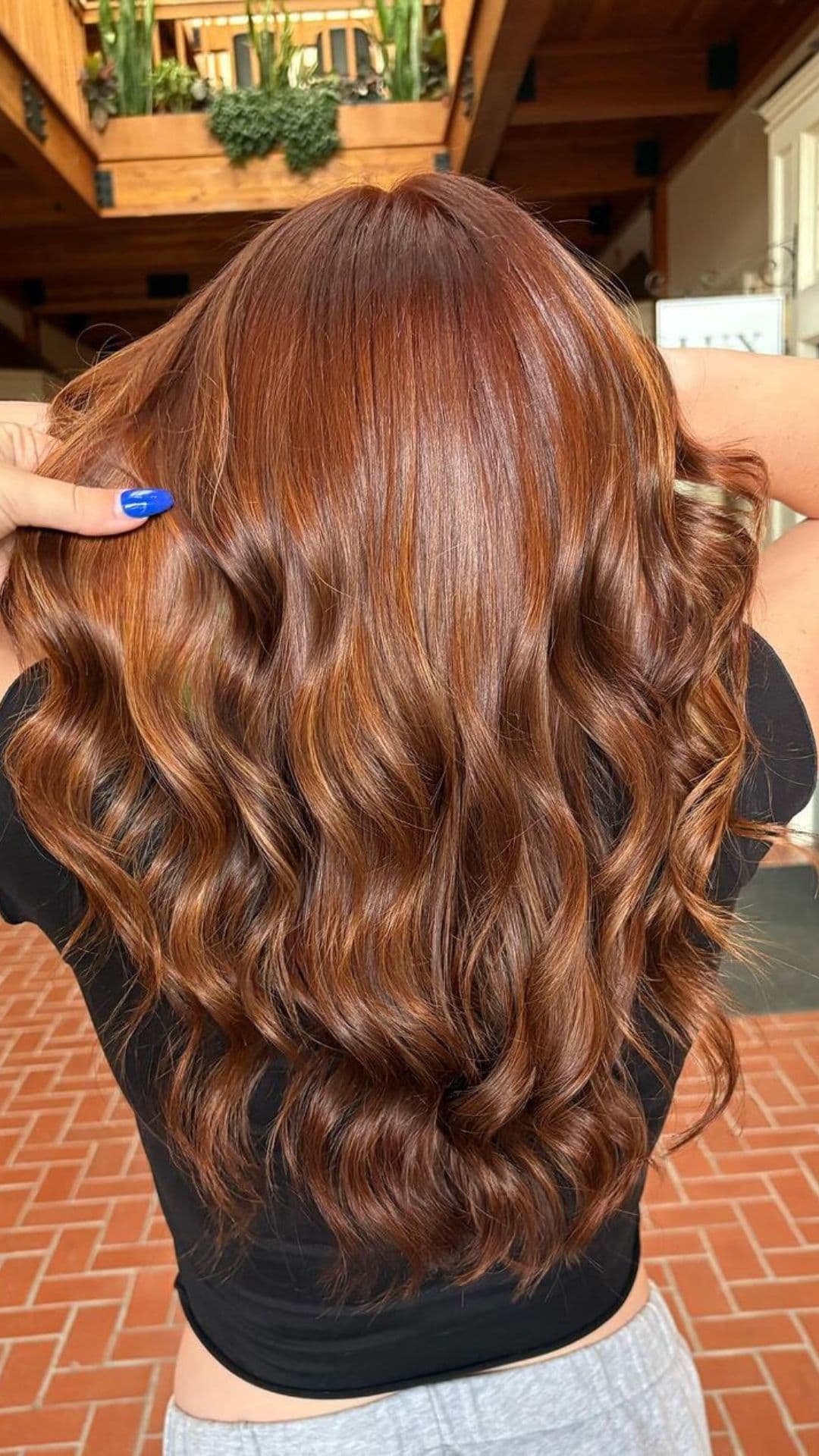 A person showcasing a styled Auburn hairstyle, with long, wavy hair, holding it gently with one hand adorned with blue nail polish.