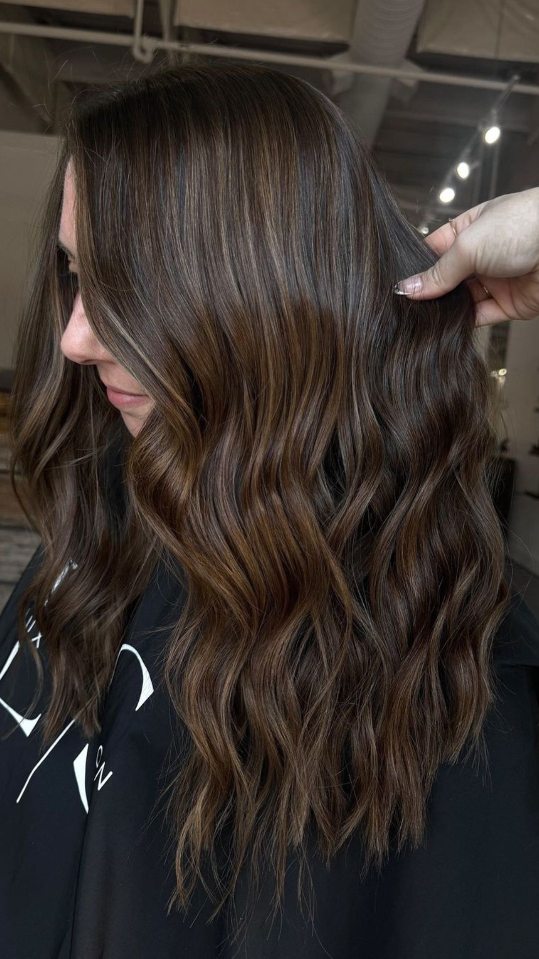 A person showcasing a wavy caramel balayage hairstyle, with a close-up view of the cascading hair highlighted against a dark background.