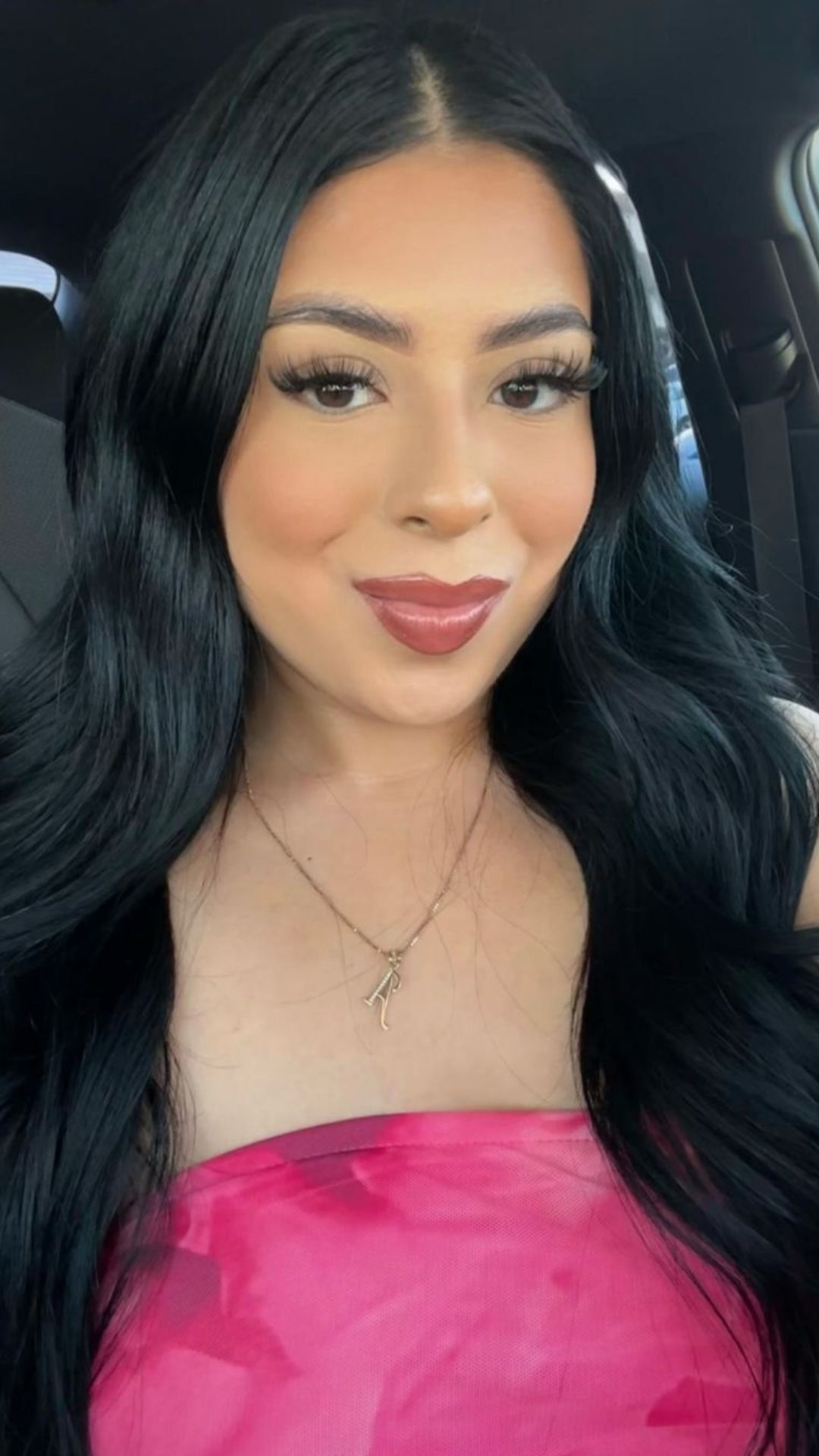 A person modeling a jet black hairstyle with long, sleek hair, wearing a strapless pink top and a necklace, sitting in a car.