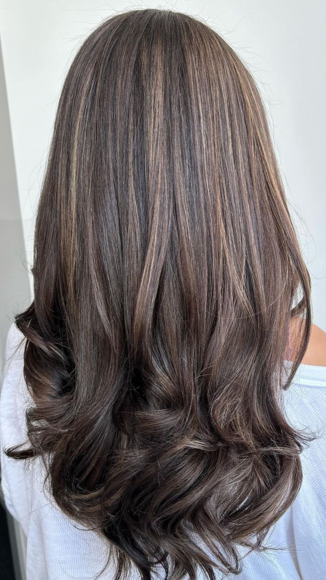 A person with long Mushroom Brown hair, featuring soft curls and subtle highlights, viewed from the back.